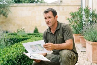 Paysagiste professionnel examinant un plan d'aménagement paysager dans un jardin de style bordelais avec haies taillées et allées en gravier